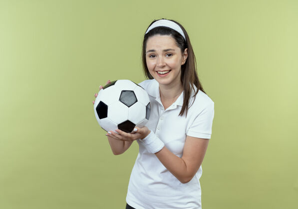 轻戴着头巾 手持足球 面带微笑的年轻健身女士站在轻盈的墙上欢呼年轻微笑