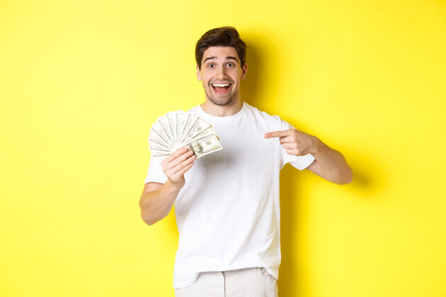 男人快乐的帅哥指着钱 信贷的概念 站在黄色的背景上成年人模特自信