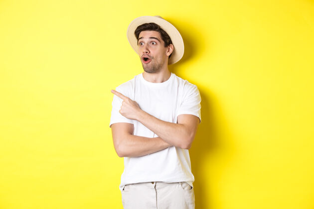 年轻旅游和生活方式的概念穿着草帽的帅哥兴奋地看了看广告 指着左上角的标志 黄色的背景帽子模特工作室