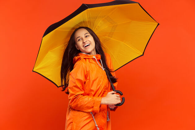 下雨那个快乐微笑的小女孩穿着秋天的橘色夹克在摄影棚摆姿势 孤立在橘色的墙上年轻冻结立场