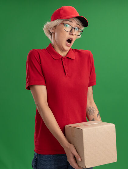 惊喜年轻的送货员身穿红色制服 戴着帽子 戴着眼镜 手里拿着一个纸板箱 站在绿色的墙上 看上去既惊讶又惊讶帽子穿上送货