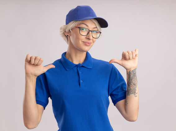 戴着身穿蓝色制服 头戴眼镜 站在白墙上用大拇指指着自己的年轻女送货员她自己帽子站着