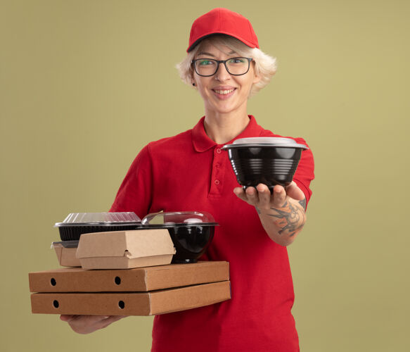 欢呼身穿红色制服 戴着眼镜 手持比萨饼盒和食品包的年轻送货员微笑着 站在绿色墙壁上的盒子盒子送货拿着