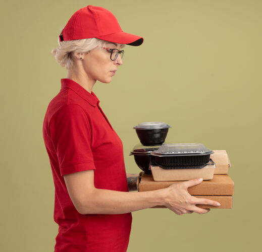 严肃年轻的送货员身穿红色制服 戴着帽子 戴着眼镜 手里拿着披萨盒和食品包 侧着身子站着 严肃的脸俯视着绿色的墙壁帽子送货侧着
