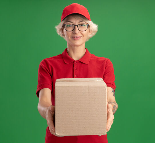 交货年轻的送货员身穿红色制服 戴着帽子 戴着眼镜 手里拿着纸板箱 站在绿色的墙边 面带微笑 自信满满立场穿女人