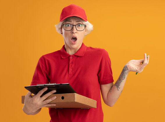 剪贴板身穿红色制服 戴着眼镜 拿着披萨盒和铅笔的剪贴板的年轻送货员站在橙色的墙上 既惊讶又惊讶年轻人铅笔帽子