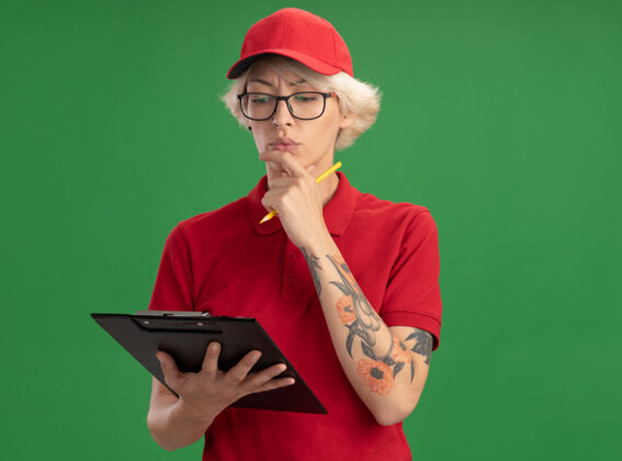 交货身穿红色制服 戴着帽子 戴着眼镜 拿着写字板和铅笔 表情严肃 站在绿色的墙上思考的年轻女送货员脸年轻立场