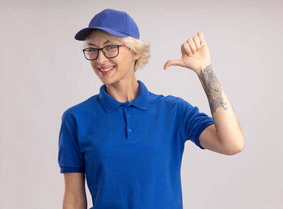 年轻人身穿蓝色制服 头戴帽子的年轻送货员面带微笑 用拇指指着站在白墙上的自己制服指着她自己