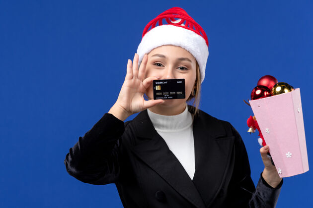 人前视图：带着圣诞树玩具和银行卡的年轻女士在蓝墙新年假期帽子年轻女士视图