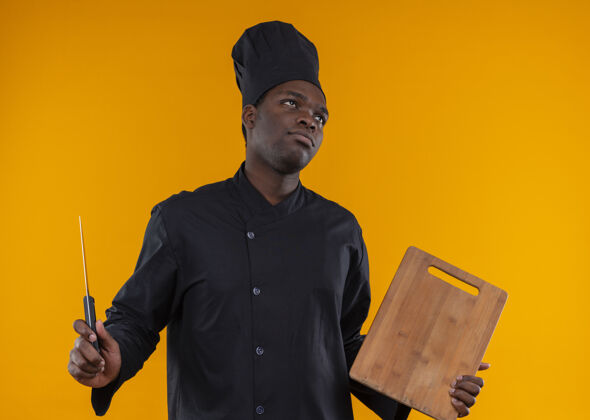 复制年轻恼怒的非洲裔美国厨师穿着厨师制服 拿着刀和砧板 看着橙色空间里的一边隔离着复制空间看壁板橙色