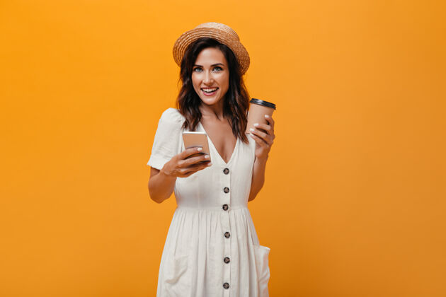 橙色背景戴草帽的黑发女孩拿着智能手机和一杯咖啡一个卷发的女人手里拿着手机和一杯茶看着相机休闲女孩沉思