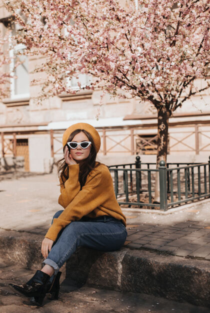 时尚的女人穿着牛仔裤和毛衣的女人在樱花的背景下靠在路边休息穿着巴黎风格的时髦服装的女人享受春天的天气的肖像美女贝雷帽温柔的花
