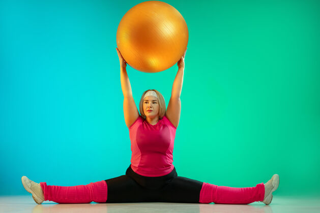 女性年轻的白人大号女模特在霓虹灯下的渐变绿色背景下训练用健身球做运动运动的概念 健康的生活方式 积极的身体 平等健康举重体重