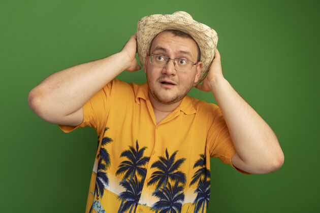 惊喜戴着橘色衬衫和夏令帽的戴眼镜的男人一边看一边惊讶地摸着他站在绿墙上的帽子站着眼镜帽子