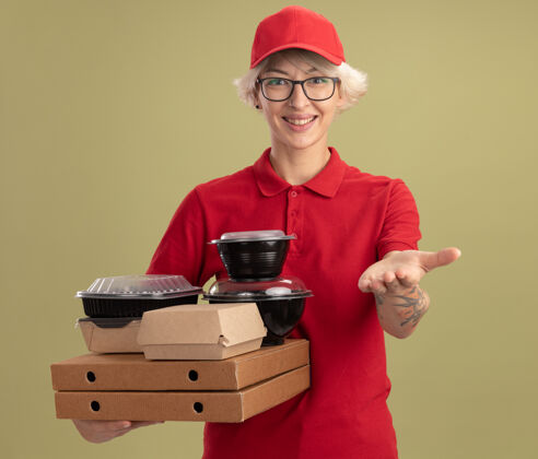 帽子身穿红色制服 戴着眼镜 手持比萨饼盒和食品包装的年轻送货员友好地站在绿色墙壁上微笑着女人食物站着