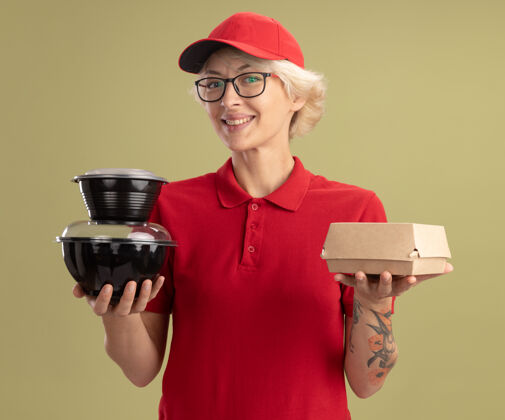 抱着年轻的送货员身穿红色制服 戴着眼镜 手里拿着食品包 自信地站在绿色的墙上帽子女人眼镜