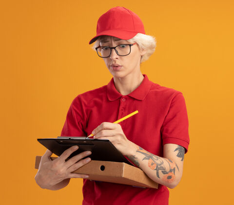 年轻身穿红色制服 戴着眼镜 拿着披萨盒 站在橘色墙上 在剪贴板上写着什么 看上去很困惑的年轻女送货员眼镜困惑女人