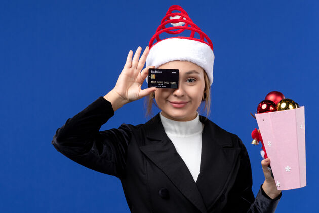 女人前视图：带着圣诞树玩具和银行卡的年轻女士在蓝墙新年情感假期年轻的女士专业圣诞节