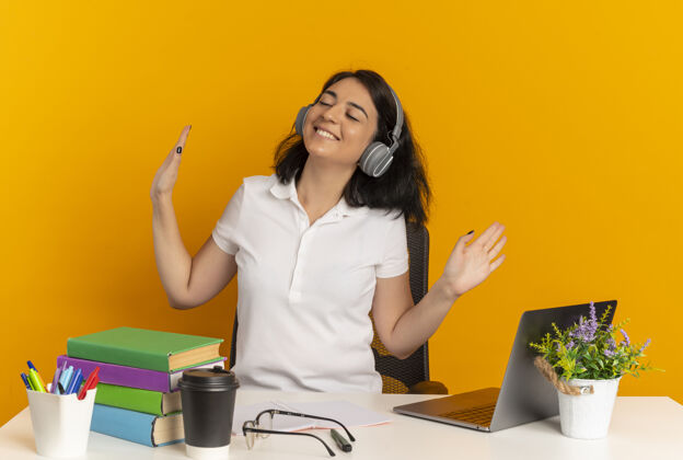 女学生戴着耳机的年轻漂亮的白人女学生坐在书桌旁 手举着学习工具 手举着 在橙色的空间和复印空间隔离开来手年轻耳机