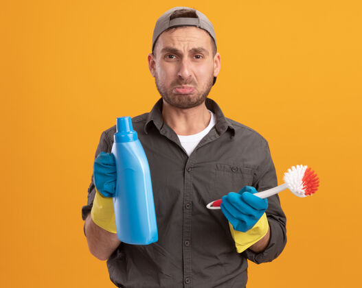年轻年轻的清洁工穿着便服 戴着橡胶手套 戴着帽子 手里拿着刷子和瓶子 手里拿着清洁用品 站在橘色的墙上 神情悲伤帽子刷子清洁