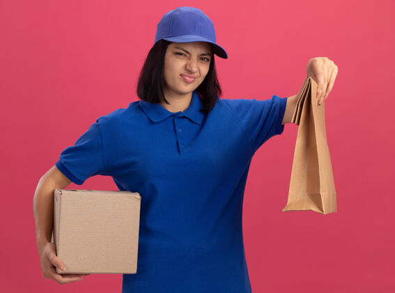 做着不满意的年轻送货女孩 穿着蓝色制服 戴着帽子 手里拿着纸包和纸板箱 站在粉红色的墙上扭着嘴制服年轻不满意