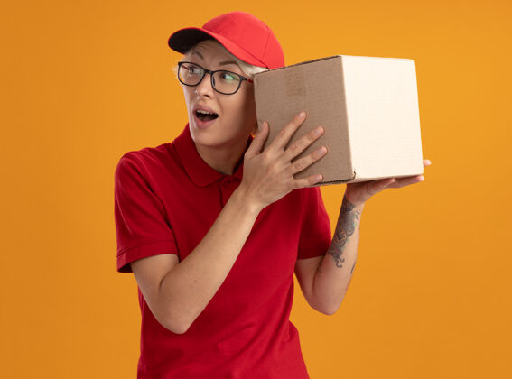 制服身穿红色制服 戴着眼镜 戴着帽子的年轻送货员站在橙色的墙上微笑着 耳朵上挂着一个纸板箱眼镜送货戴着