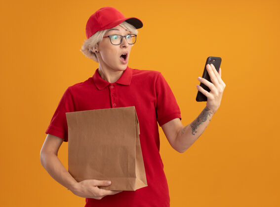 帽子年轻的送货员身穿红色制服 戴着帽子 手里拿着一个纸包 站在橙色的墙上 看着自己的智能手机 感到困惑和惊讶女人惊喜年轻