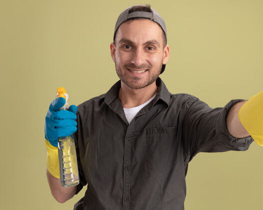 帽子年轻的清洁工穿着休闲服 戴着橡胶手套 手里拿着喷雾瓶 站在绿色的墙壁上 面带微笑穿举行清洁