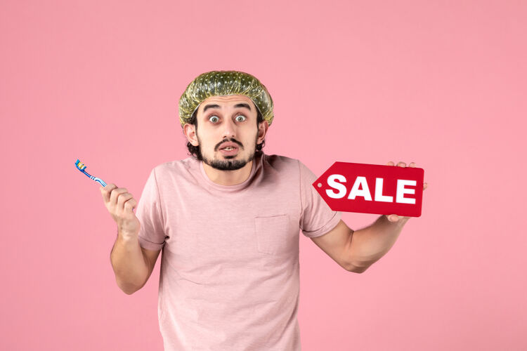 销售正面图年轻的男性正在清洁他的牙齿 拿着粉红色背景上的销售铭牌牙刷脸帽子