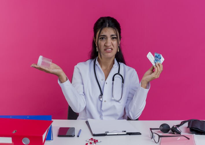 女穿着医用长袍和听诊器的年轻女医生坐在办公桌旁 手里拿着医疗工具 手里拿着隔离在粉红色墙上的药品和烧杯年轻长袍穿