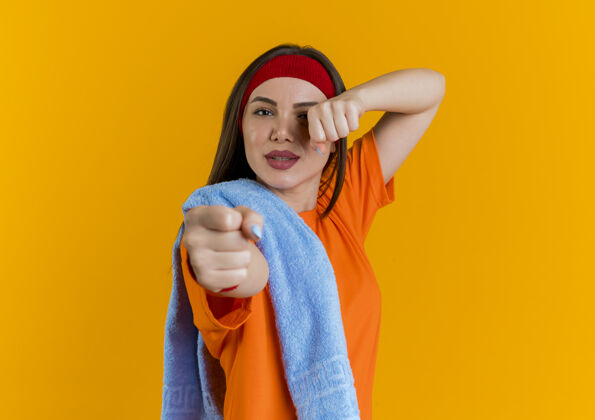 橙色自信的年轻运动女性戴着头带和手镯 肩上拿着毛巾 做着拳击动作 隔离在橙色的墙上 留着复制空间年轻头带佩戴