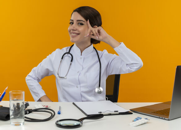 触摸面带微笑的年轻女医生穿着医用长袍和听诊器坐在办公桌旁 手持医疗工具和笔记本电脑 手指孤立在黄色墙壁上触摸头部工具笔记本电脑医生