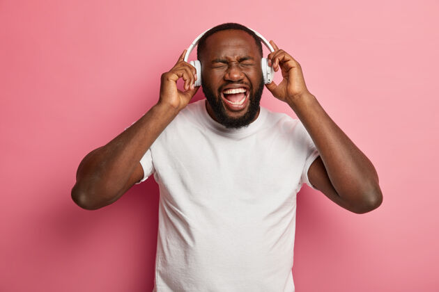 室内情绪轻松无忧无虑的胡须男人听最喜欢的歌曲从播放列表 娱乐室内 戴着白色耳机的耳朵 休闲t恤 在粉红色的墙上摆姿势成功人人