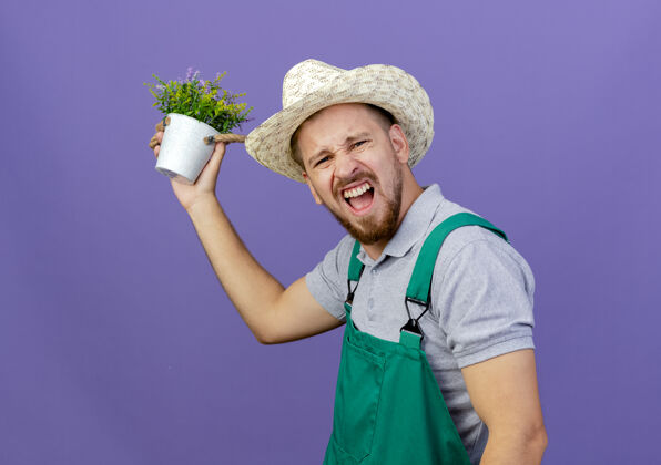 造型自信的年轻英俊的斯拉夫园丁 身着制服 戴着帽子站在侧视图中 手里拿着花盆 看起来孤立地站在紫色的墙上 有复制空间花园站立风景