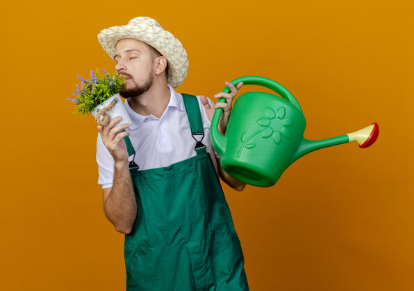 嗅年轻英俊的斯拉夫园丁穿着制服 戴着帽子 拿着花盆和浇水罐 闭着眼睛在橙色的墙上嗅着花封闭帅气浇水