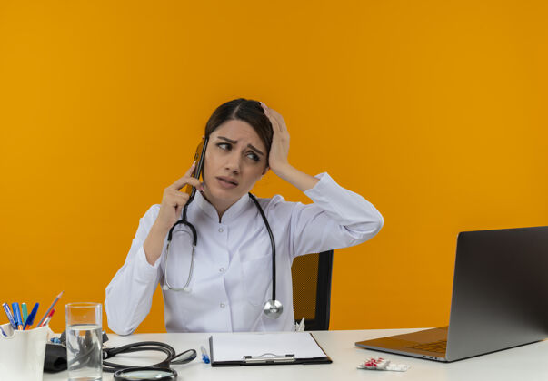 电话失望的年轻女医生穿着医用长袍和听诊器坐在办公桌旁 拿着医疗工具和笔记本电脑打电话 看着隔离在黄色墙上的一面医疗笔记本电脑女性