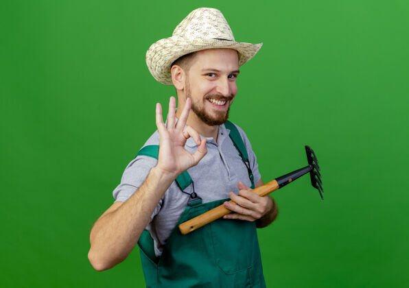 好面带微笑的年轻英俊的斯拉夫园丁 穿着制服 戴着帽子 手里拿着耙子 看上去正在做一件好事 绿色的墙上孤立着一块牌子帽子微笑斯拉夫