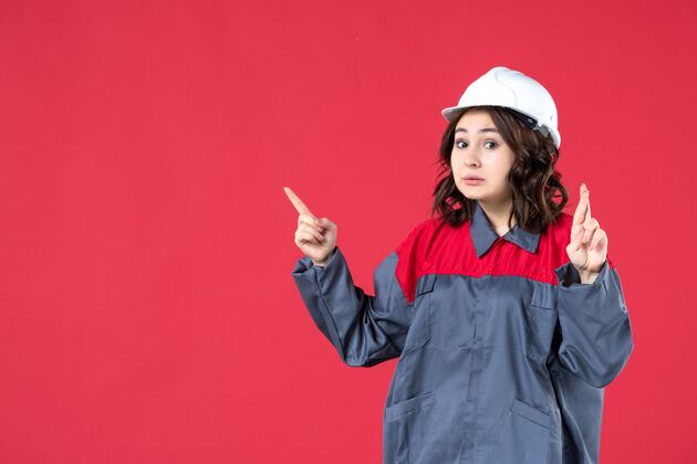 微笑正面近距离观看穿着制服 戴着安全帽 在孤立的红墙上交叉手指做出“叫我”的手势的惊讶的女建筑工人成人人帽子