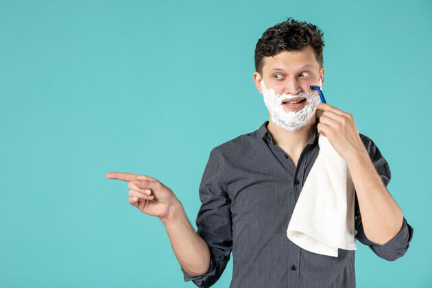 胡子正面图：年轻的男性用剃刀在蓝色背景上刮泡沫脸前面剃须脸