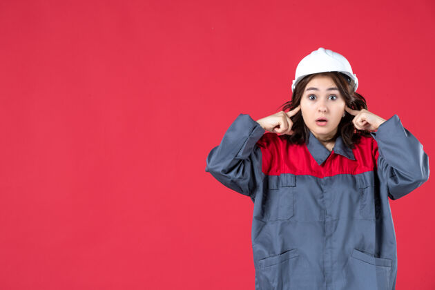 微笑特写镜头中惊讶的女建筑工人穿着制服 戴着安全帽 在孤立的红墙上闭着耳朵人硬人