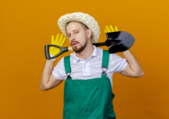 斯拉夫自信的年轻英俊的斯拉夫园丁 穿着制服 戴着帽子和园艺手套 脖子后面拿着铁锹 隔离在橙色的墙上 留着复制空间帽子手套制服