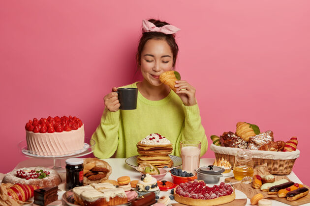 煎饼满足的亚洲女人每天每顿饭都吃美味的牛角包 喝茶 在喜庆的餐桌上摆姿势 沉迷于甜食 在粉色背景下摆姿势果酱美味微笑