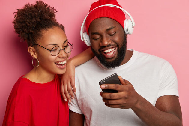 青春快乐的少数民族妇女和男子在智能手机上观看有趣的视频 戴着红帽子和白t恤的黑人男子 戴着耳机 向女友展示新的应用程序高兴歌曲手机