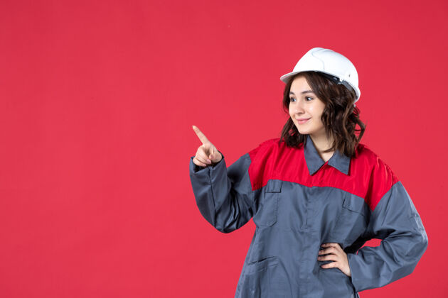 模特微笑的女建筑工人的特写镜头穿着制服 戴着安全帽 指着孤立的红墙上的东西漂亮尖肖像