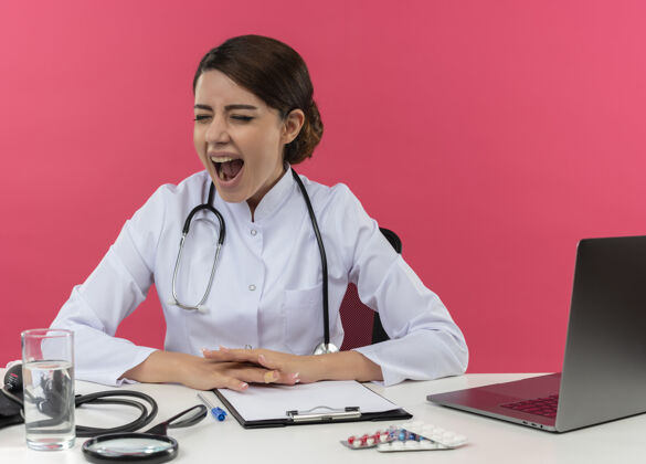 办公桌年轻的女医生穿着医用长袍和听诊器坐在办公桌旁 手里拿着医疗工具和笔记本电脑 双手放在办公桌上 闭着眼睛尖叫着站在粉红色的墙上工具女年轻