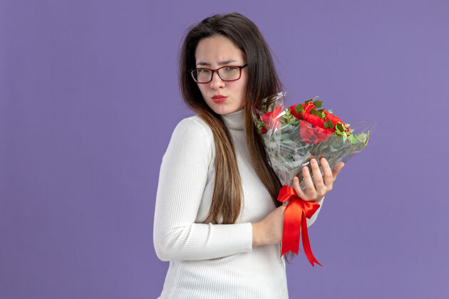 年轻穿着休闲服的年轻美女手持一束红玫瑰 带着怀疑的表情看着镜头站在紫色背景下的情人节概念怀疑站着天