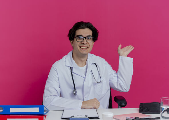 手面带微笑的年轻男医生 穿着医用长袍 戴着听诊器 戴着眼镜 坐在办公桌旁 手里拿着医疗工具 粉红色的墙上孤立着一只空手长袍微笑年轻
