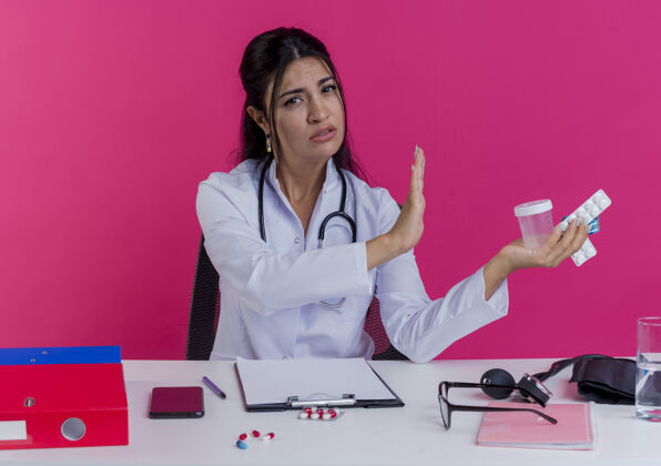长袍年轻的女医生穿着医用长袍和听诊器 坐在办公桌旁 手里拿着医疗工具 手里拿着药品和烧杯 一动不动地站在粉红色的墙上坐着女药物