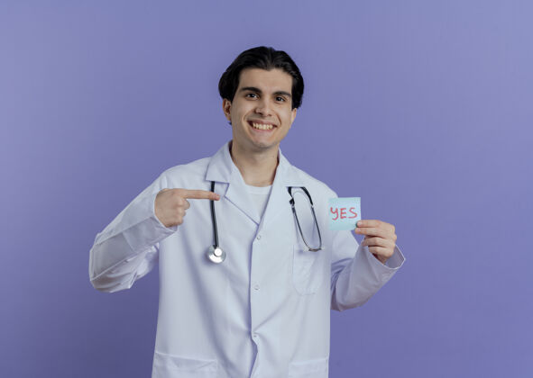 微笑面带微笑的年轻男医生穿着医用长袍 手持听诊器 显示“是”字条 指着它 隔离在紫色的墙上 留有复印空间男性穿听诊器
