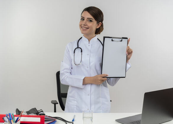 工具微笑着的年轻女医生穿着医用长袍 听诊器站在桌子后面 手持医疗工具和笔记本电脑 将剪贴板隔离在白色的墙上医生微笑穿